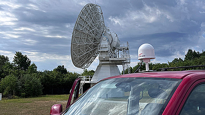 A large satellite dish in an open field with a red vehicle in the foreground. The vehicle is equipped with a small white dome-shaped antenna. Dark clouds are visible in the sky, suggesting incoming weather changes.