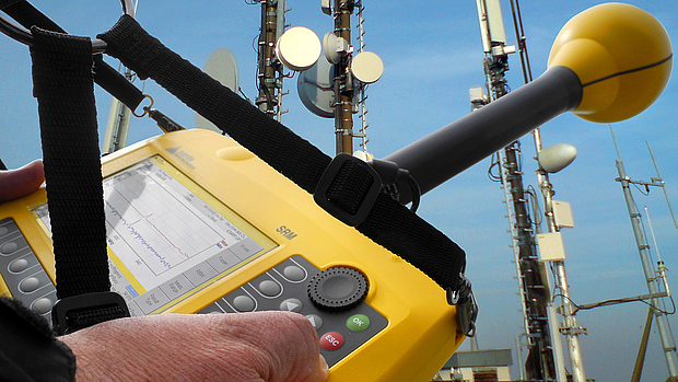 Acceptance Testing – Ensuring System Compliance & Quality A technician using a yellow handheld spectrum analyzer to test signal quality near a communication tower, representing LS telcom’s acceptance testing services for network validation.