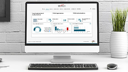 Computer Display Showing Spectrum Management Dashboard A desktop computer on a white desk displaying a spectrum management dashboard with charts and graphs. The screen shows user applications, TDMA applications, and authorizations with pie charts, bar graphs, and summary statistics. A plant in a white vase and a wireless mouse are placed next to the keyboard.
