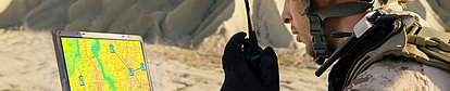 Soldier Using Radio and Topographic Map A soldier in tactical gear using a handheld radio while observing a laptop displaying a detailed topographic map. The map shows terrain features and marked points, suggesting a navigation or mission planning scenario in a desert environment.